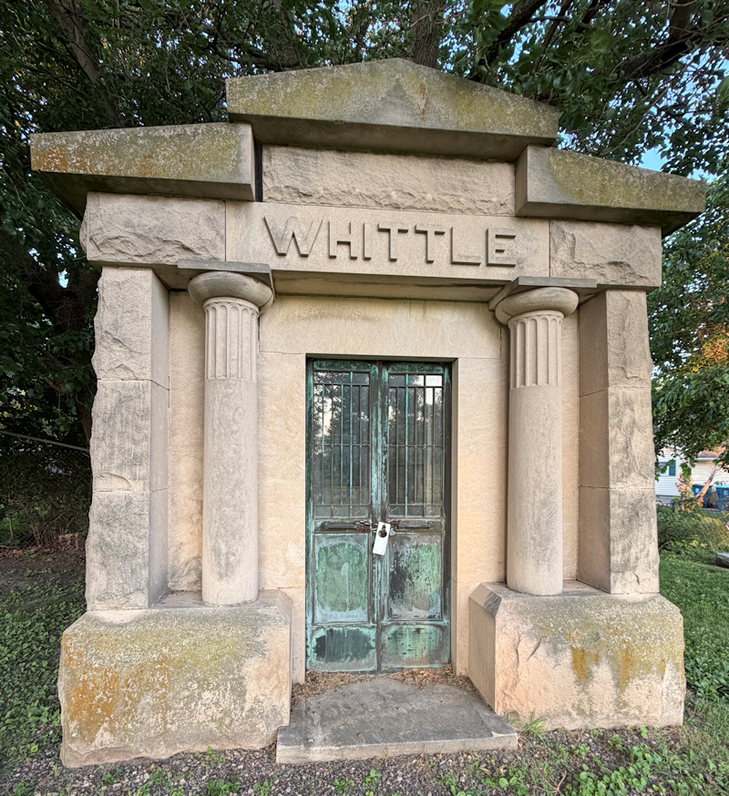 Photo of Whittle Mausoleum in Peru City Cemetery taken in 2026 in the summer. The structure is made of creme colored stone with a copper door that has turned green over time and is set back into the structure with a pad in front of it. The word "Whittle" is carved above the door.